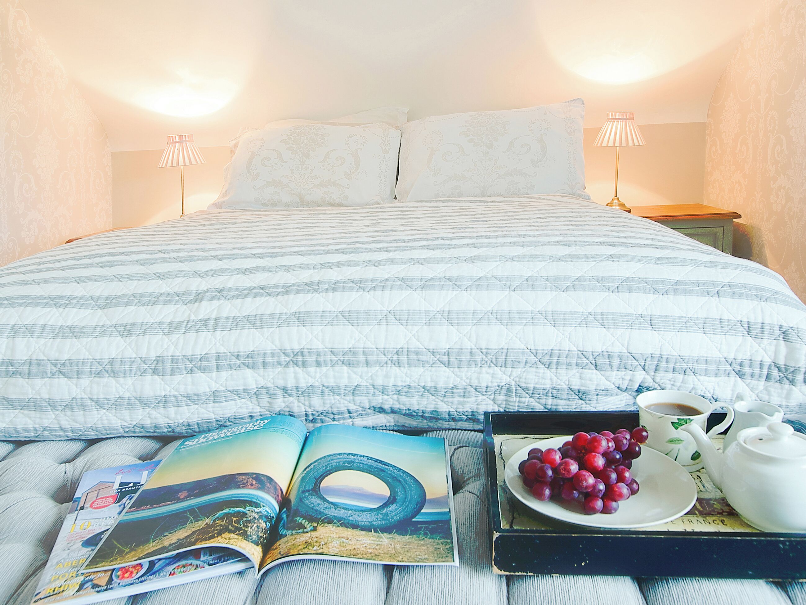 Un lit bien fait, recouvert d’une courtepointe, une table basse avec des raisins et une théière, et un magazine posé sur le lit.