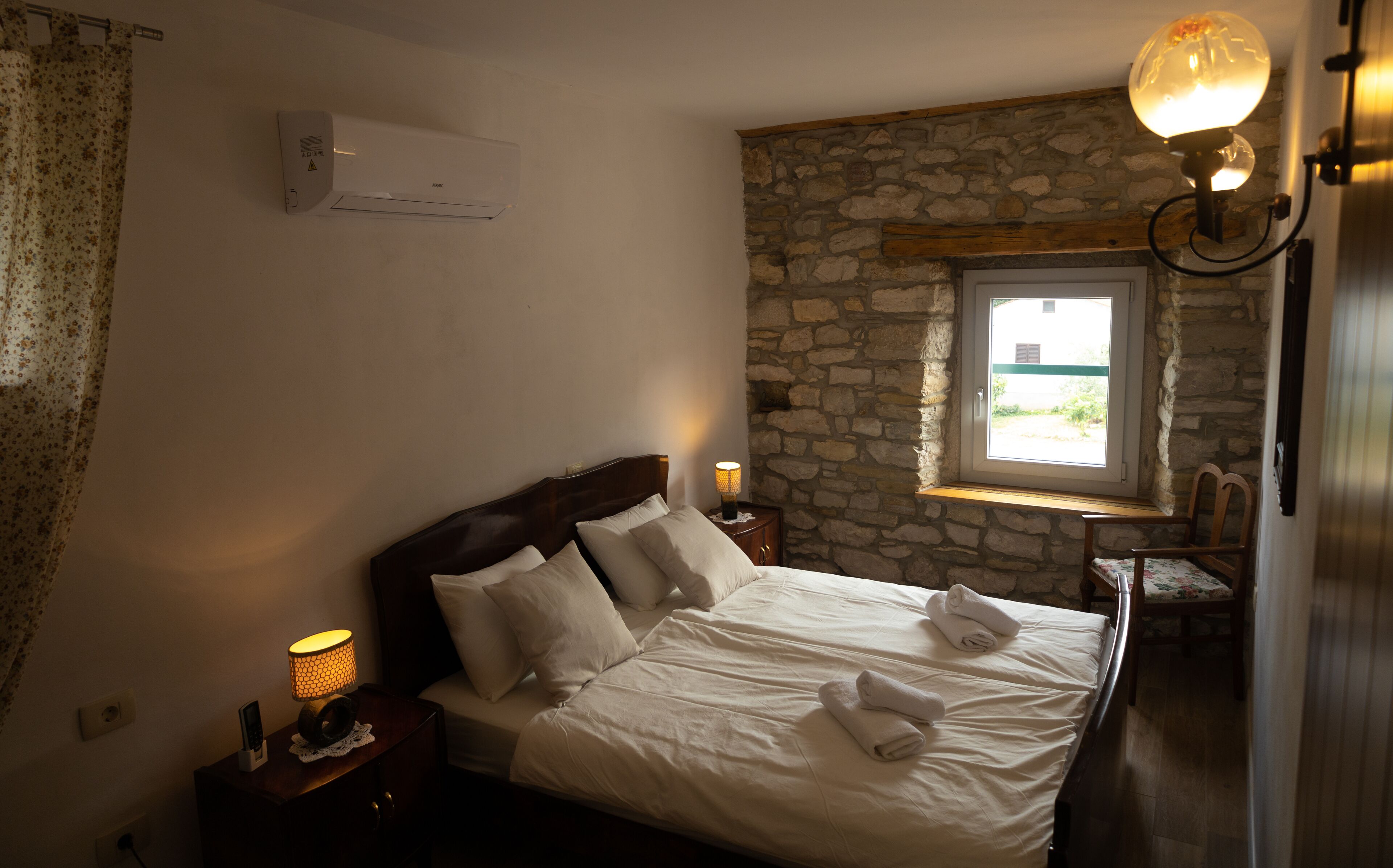 A bedroom with a stone wall, a window with a view, a bed with white linens, and a bedside lamp.