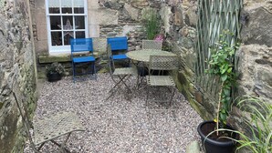 A stone-walled courtyard with a window, blue chairs, a small table, and potted plants. - 3 bedroom accommodation in Comrie, near Crieff (Comrie, near Crieff)