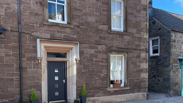 A two-story stone building with a dark-colored door, multiple windows, and a small balcony with potted plants. - 3 bedroom accommodation in Comrie, near Crieff (Comrie, near Crieff)
