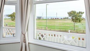 A room with two windows, a view of a grassy area, a white fence, and a building in the distance. - 5 bedroom accommodation in Paignton (Paignton)