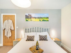 Ein Schlafzimmer mit einem Bett, zwei Nachttischen, einer Holztür, einem an der Tür hängenden Bademantel, einem gerahmten Bild an der Wand und einem Tablett mit Flasche und Glas auf dem Bett.