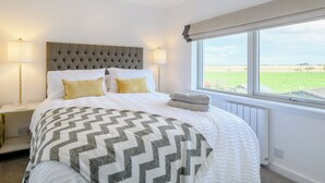 A bedroom with a large bed, two bedside lamps, a window with a view of a green field, and a radiator.