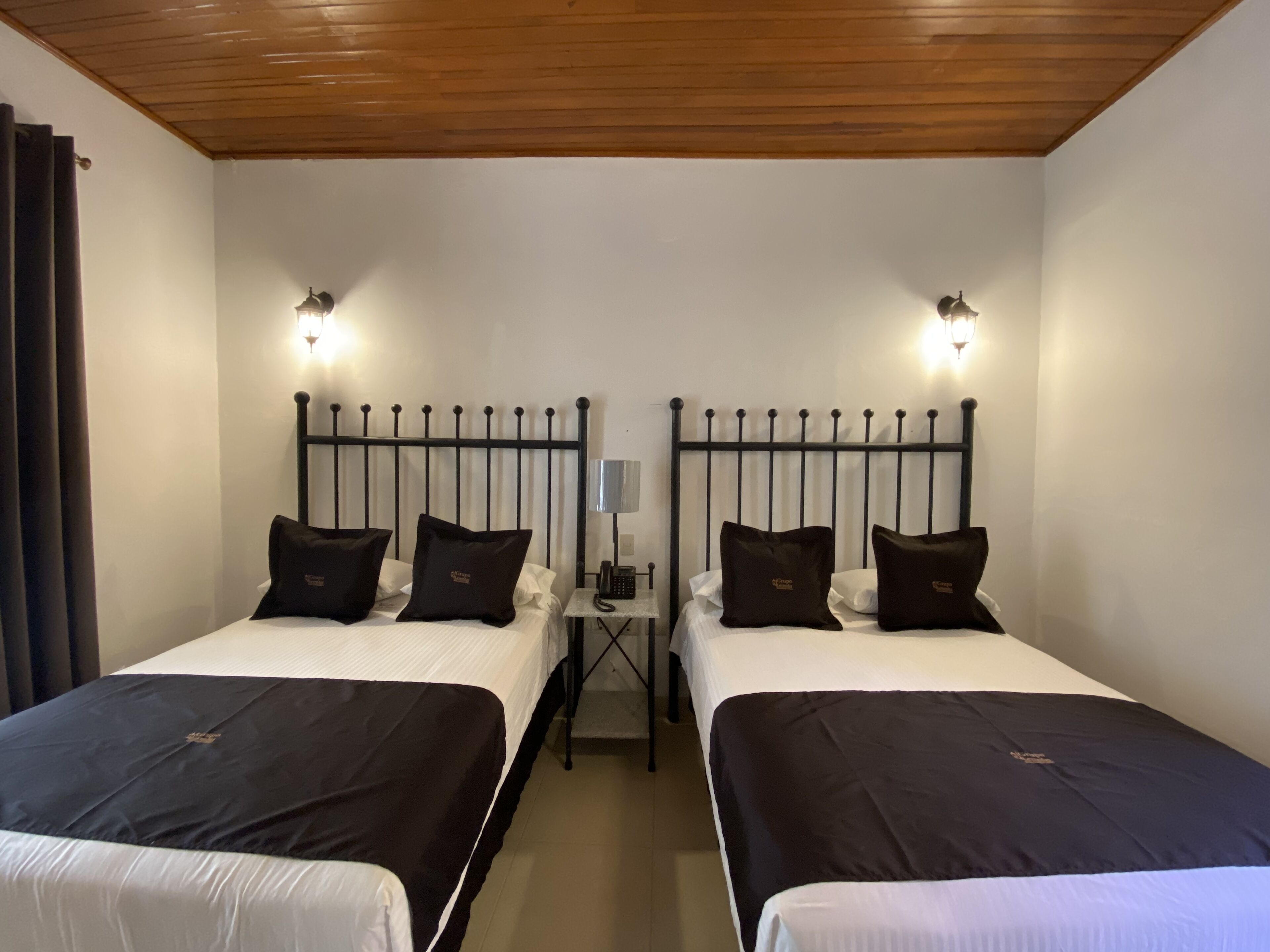 Een hotelkamer met twee bedden, elk met een zwart hoofdbord en witte beddengoed. Twee nachtkastjes met lampen en een klein tafeltje tussen de bedden. De kamer heeft een houten plafond en witte muren.