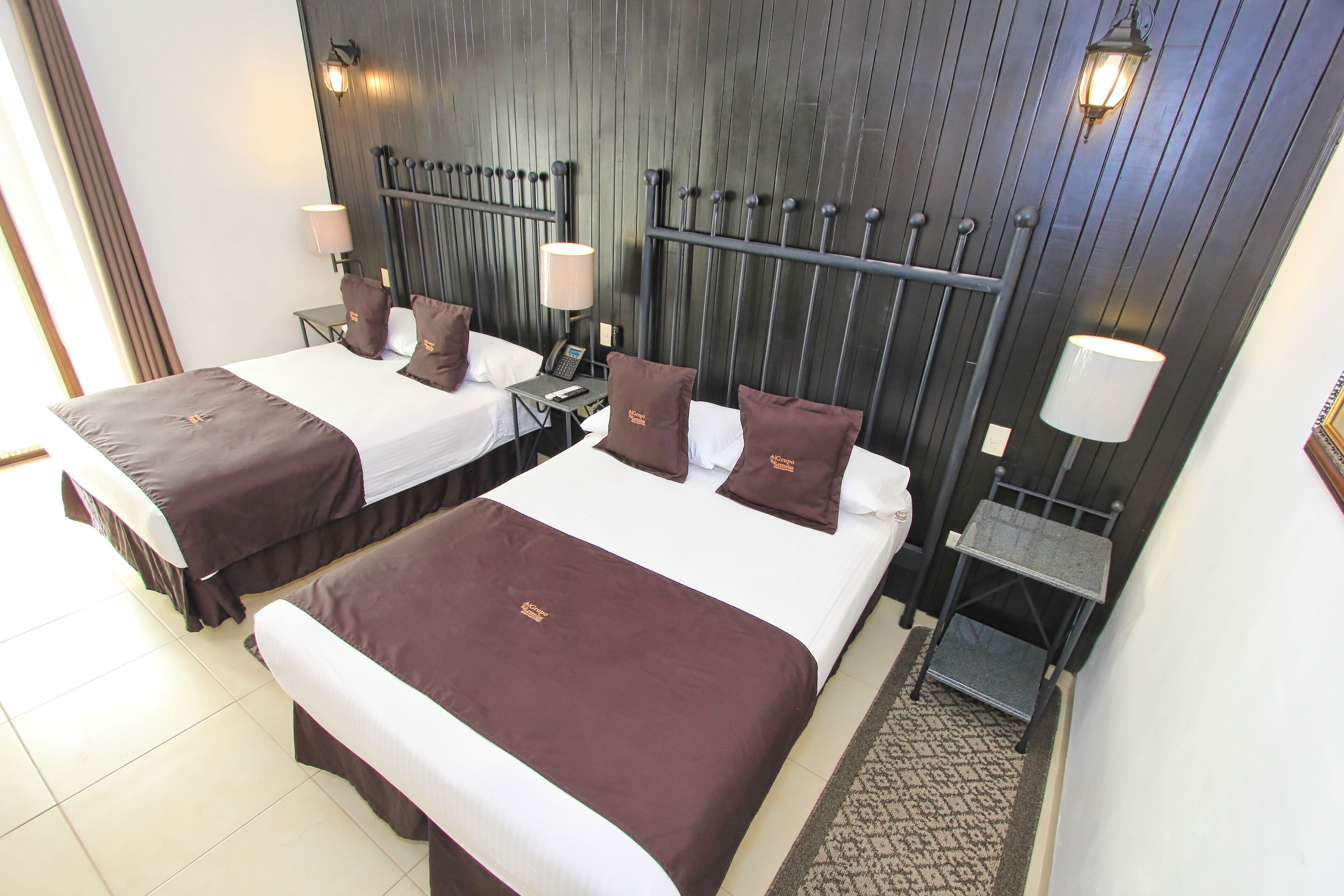 A hotel room with two beds, each with a dark brown bedspread, white linens, and matching pillows. There are bedside tables with lamps and a phone. The room has a wooden headboard and a patterned rug on the floor.