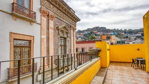 A balcony with ornate railings, a yellow wall, and a view of a cityscape in the background.