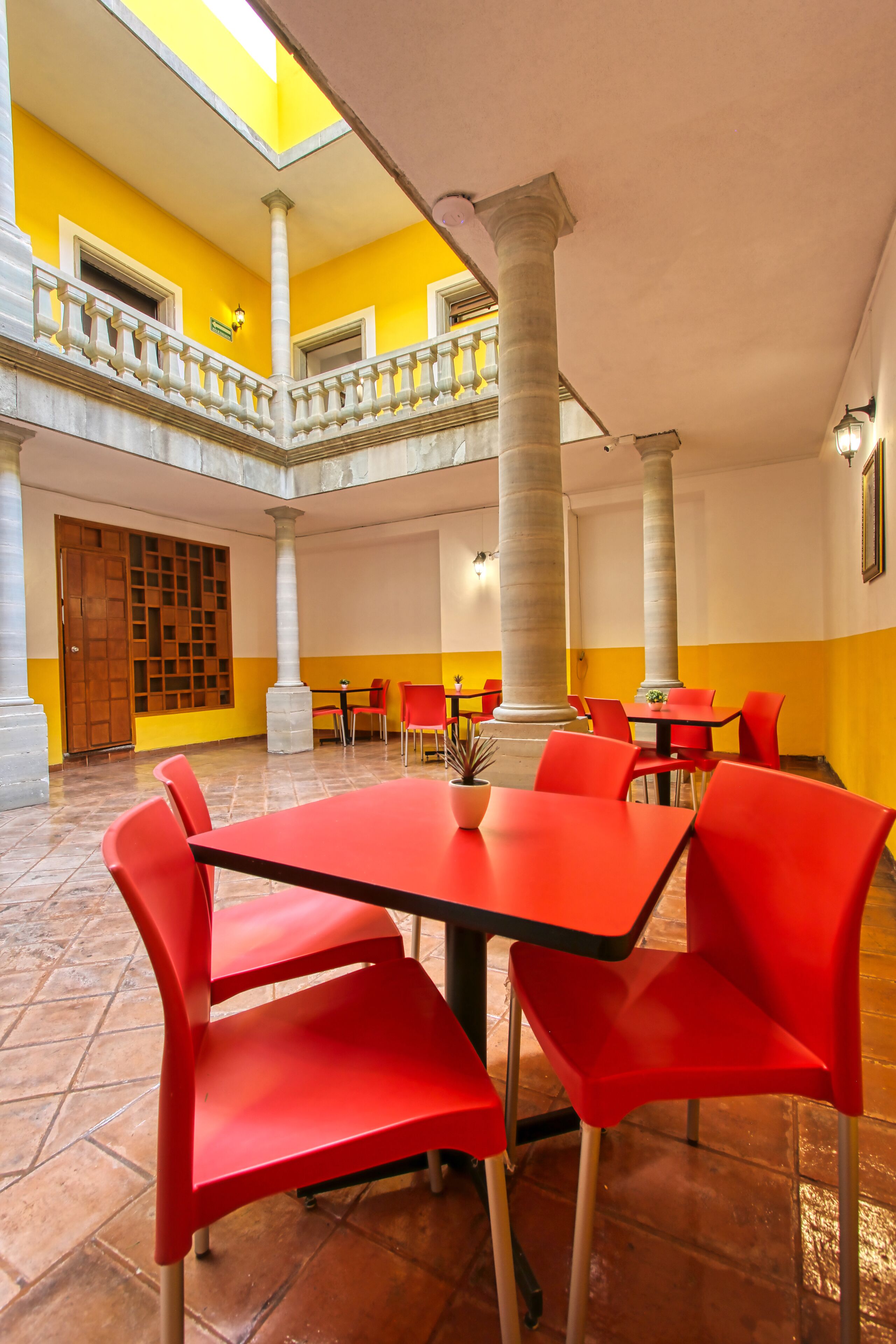 A spacious indoor area with a high ceiling, featuring a central open space surrounded by red chairs and tables, with a yellow and white color scheme and a wooden door in the background.