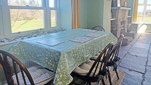 Un coin repas avec une table dressée pour quatre personnes, des chaises, une cheminée en pierre et une vue sur l’extérieur grâce à de grandes fenêtres.