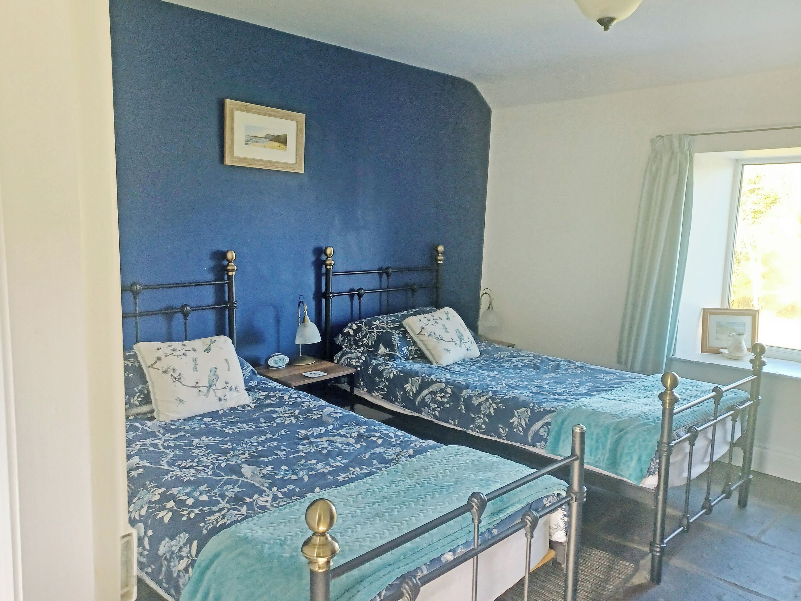 A bedroom with two beds, a bedside table with a lamp, a framed picture on the wall, and a window with curtains.