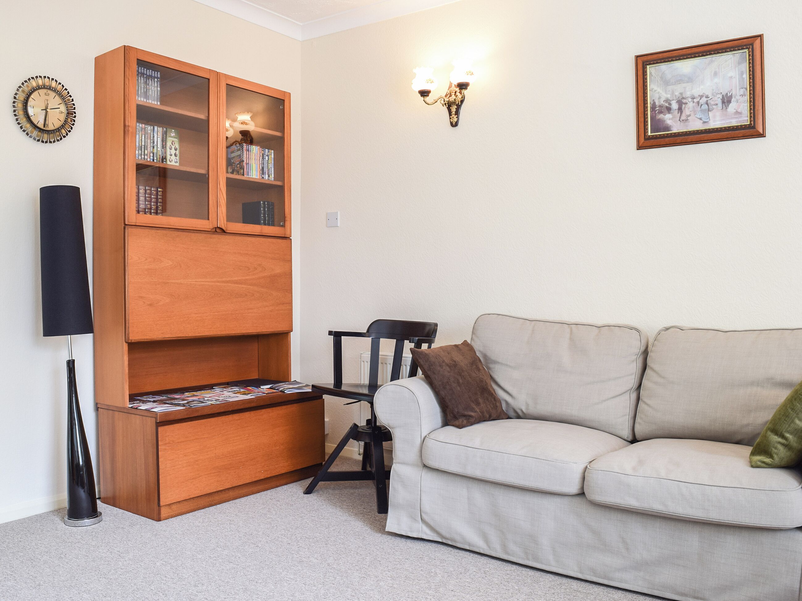 Een woonkamer met een beige bank, een houten boekenkast, een zwarte vloerlamp en een aan de muur bevestigd fotolijstje.