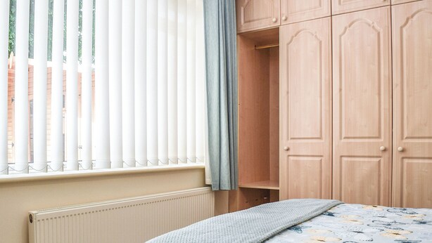 Un dormitorio con cama, radiador, ventana con persianas y armarios de madera.