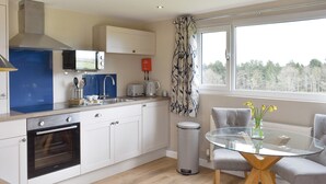 A modern kitchen with a built-in oven, a microwave, and a sink. There is a dining area with a glass table and two chairs. - Pheasant Lodge at Wood House (Consett)