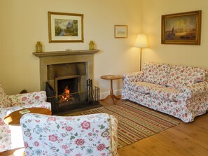 A living room with a fireplace, floral-patterned sofa, armchairs, and a small round table. - Keepers Cottage Burradon (Burradon near Thropton)
