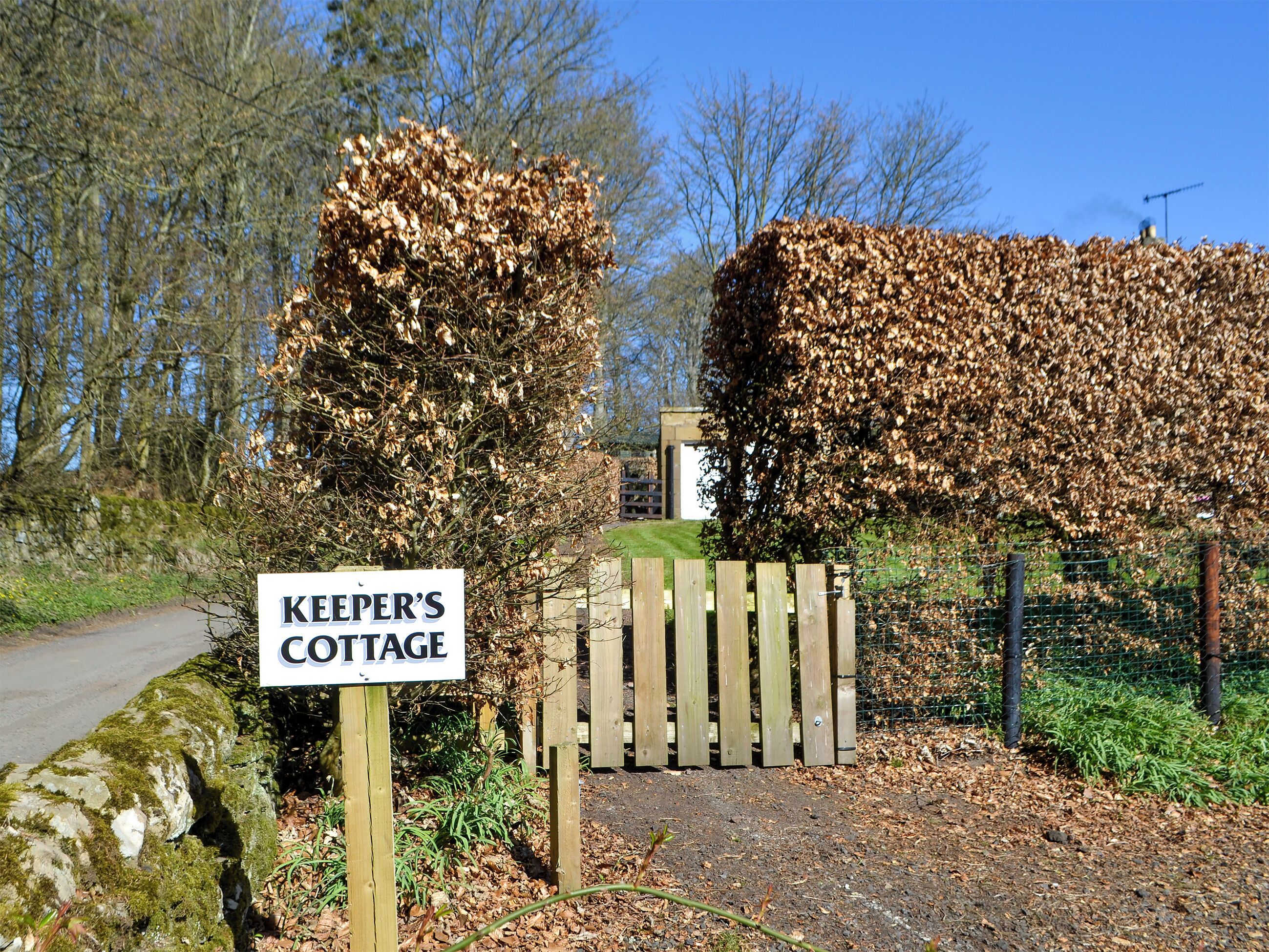 A wooden gate with a sign reading "KEEPER'S COTTAGE" in front of a pile of leaves.
