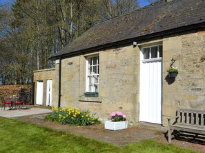 A stone building with a white door, a window with a green frame, and a bench outside. - Keepers Cottage Burradon (Burradon near Thropton)
