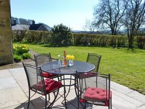 A round metal table with four chairs, each with a red cushion, set on a patio with a view of a grassy area and trees.
