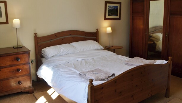 A neatly made bed with white linens, a wooden dresser with drawers, a bedside table with a lamp, and a wooden wardrobe.