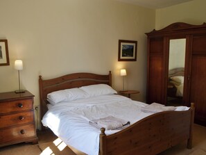 A neatly made bed with white linens, a wooden dresser with drawers, a bedside table with a lamp, and a wooden wardrobe.