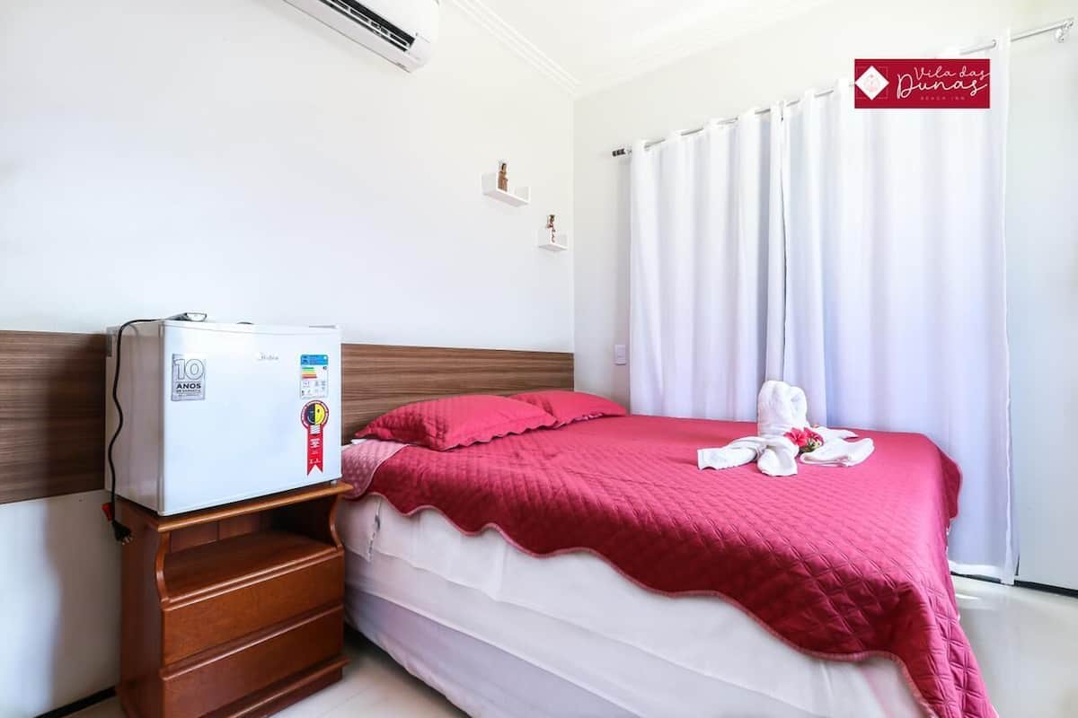 A hotel room with a bed, a small wooden nightstand, a white refrigerator, and a window with white curtains.