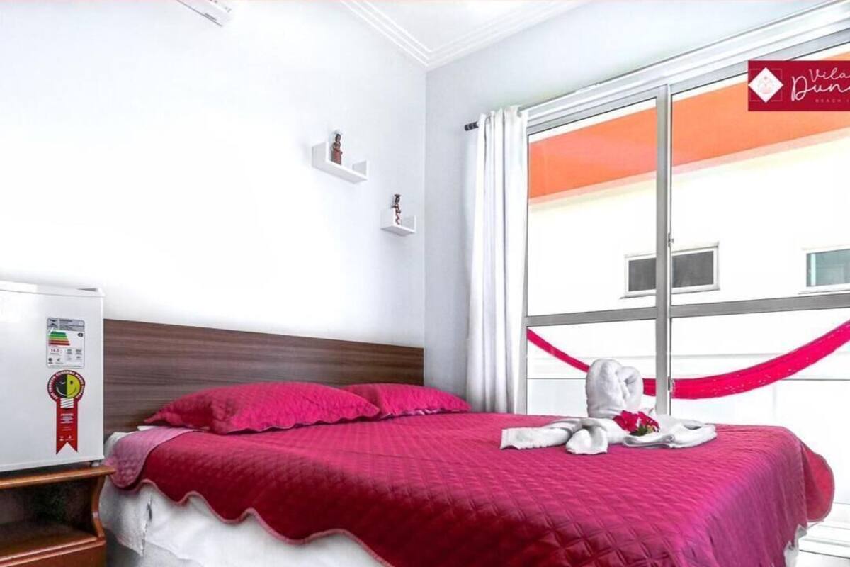 A hotel room with a bed, a nightstand, a window with curtains, and a view of a building outside.