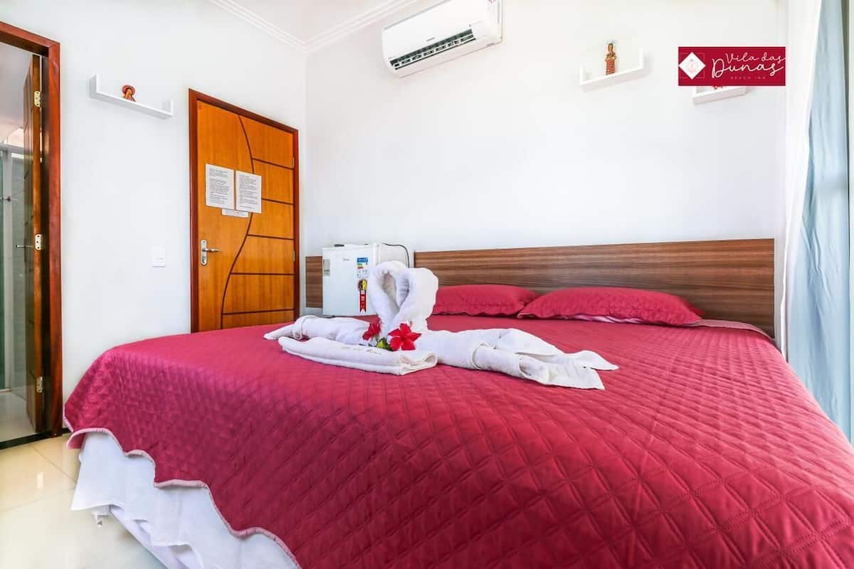 A hotel room with a red bedspread, a wooden door, a white air conditioning unit, and a sign on the wall.