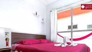 A hotel room with a bed, a nightstand, a window with curtains, and a view of the outside.
