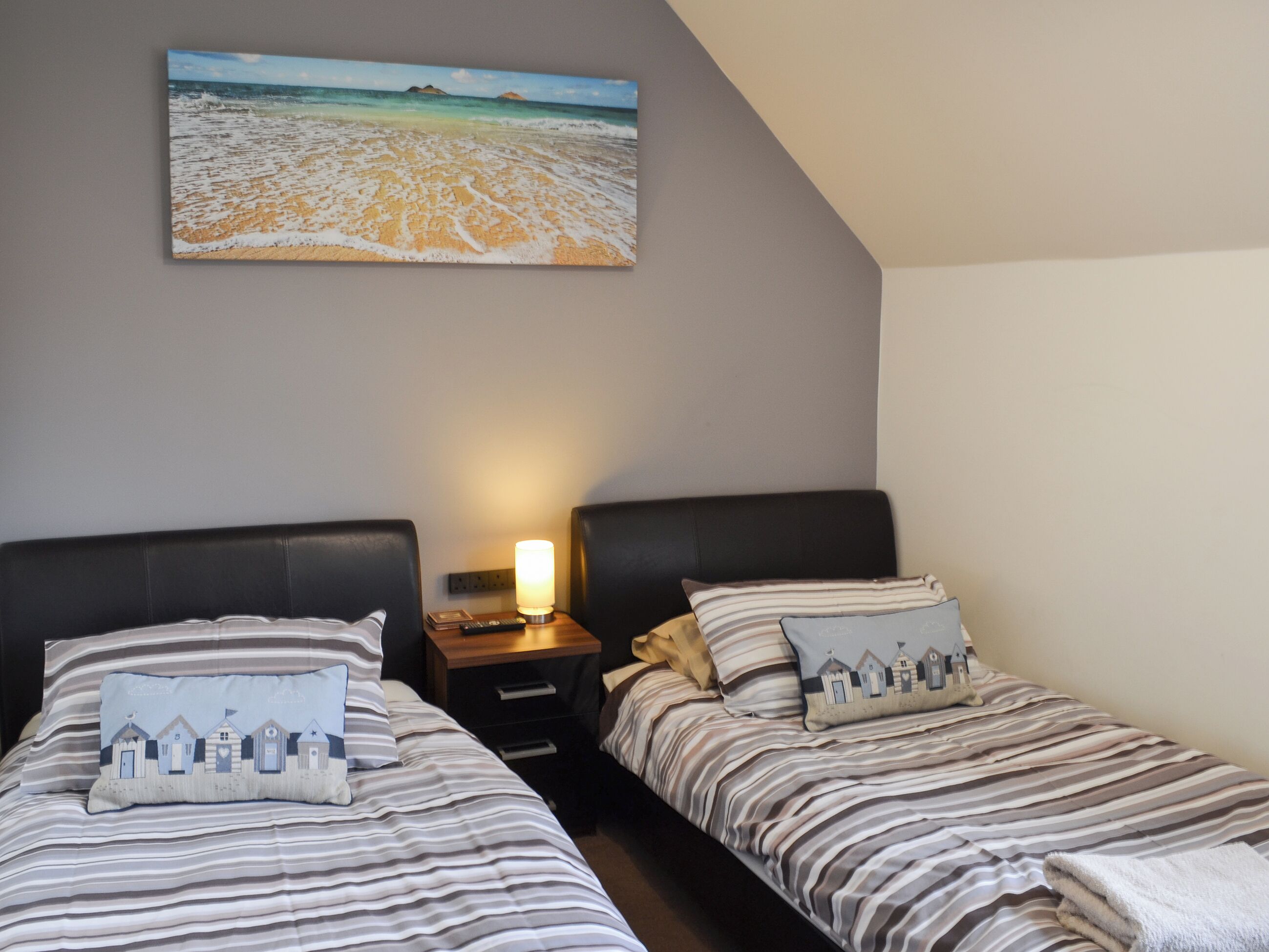 A bedroom with two beds, striped bedding, a beach-themed pillow, a bedside lamp, and a painting of a beach scene.