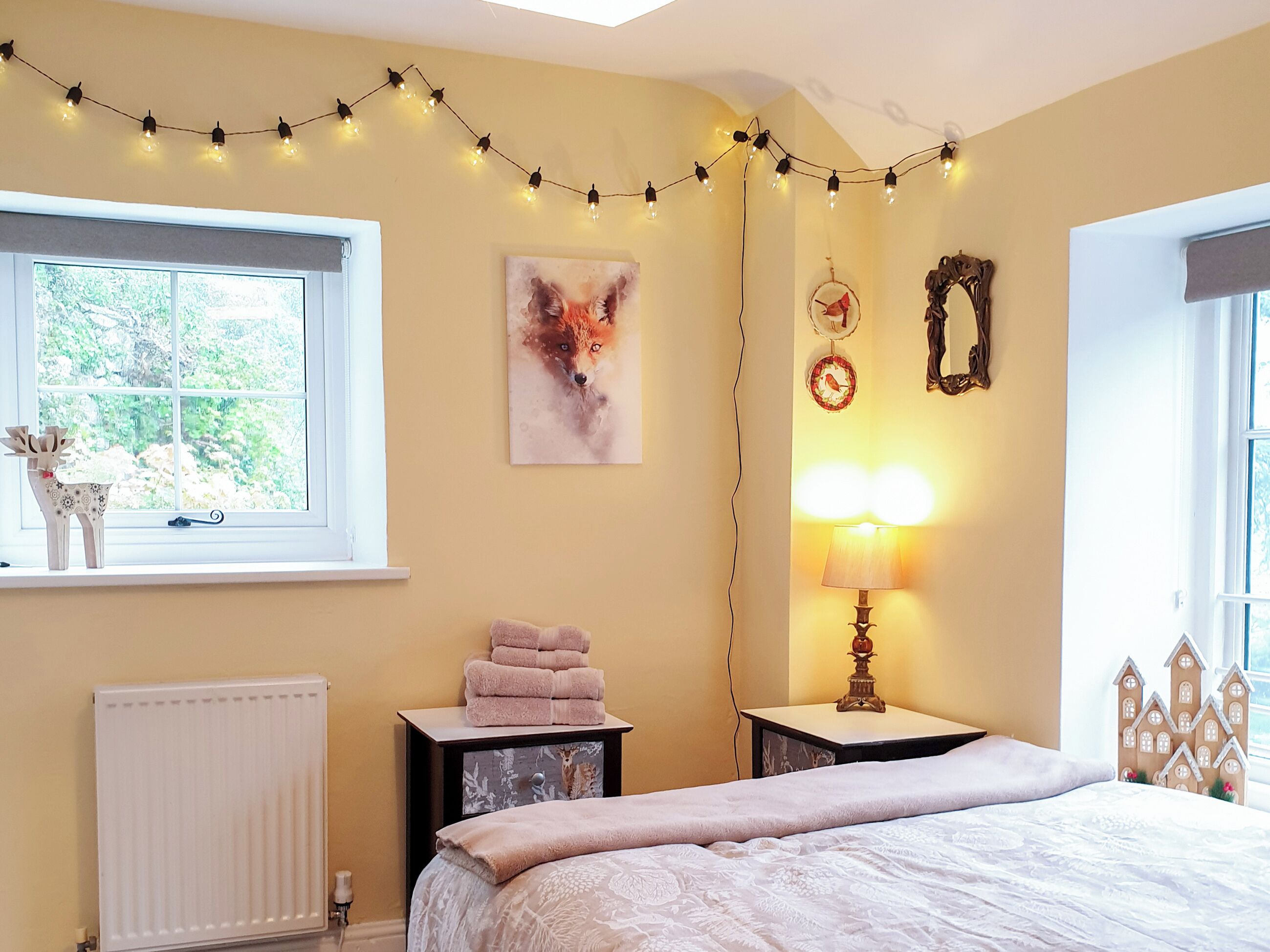A bedroom with a bed, a bedside table with a lamp, a window with curtains, a radiator, a wall with a fox painting, and string lights.
