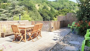 Una terraza de madera con una mesa y sillas, rodeada por un cerco de madera y vistas a una ladera boscosa.
