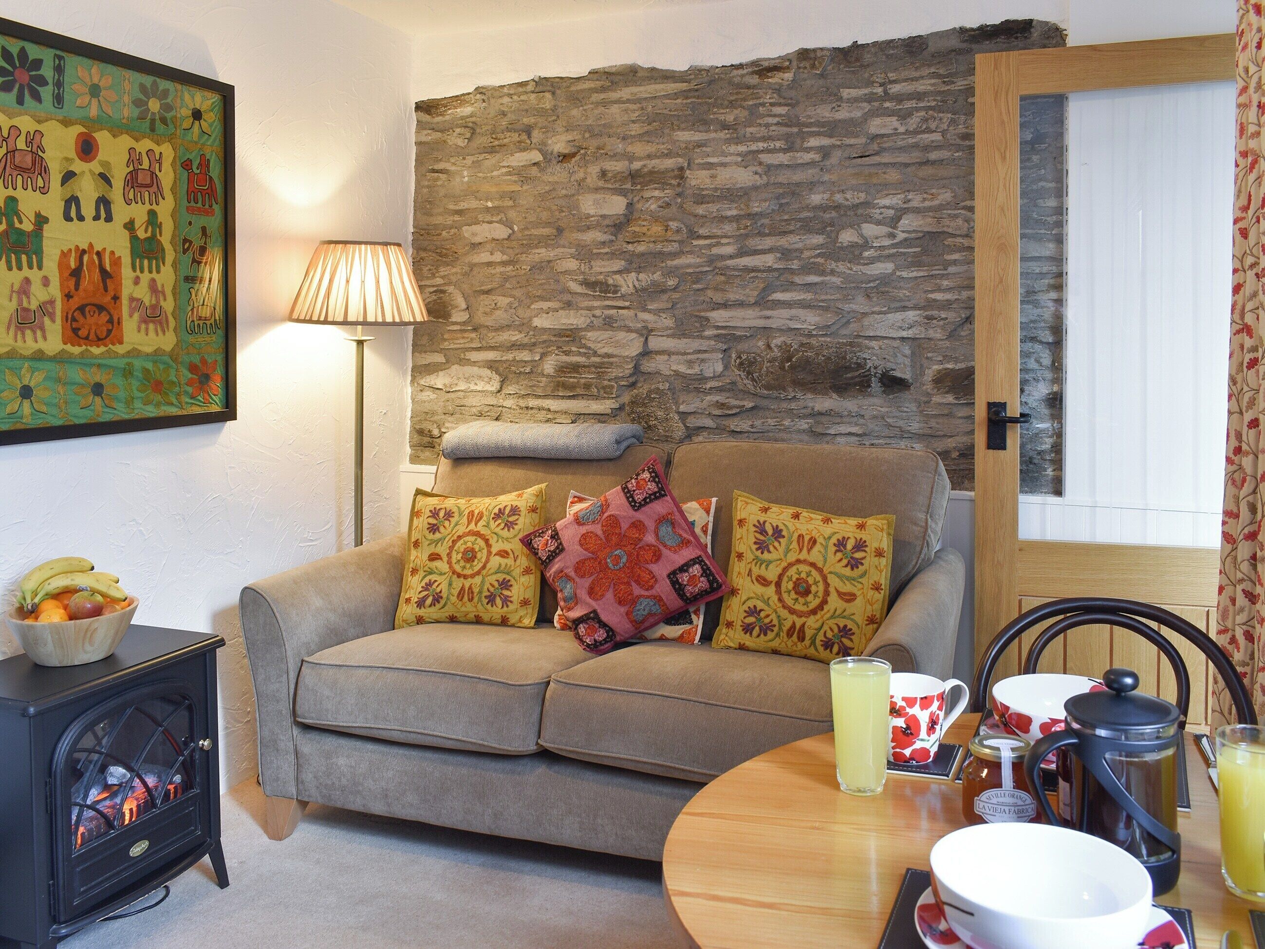 A cozy living room with a stone wall, a sofa with colorful cushions, a small black stove, and a dining table with breakfast items.
