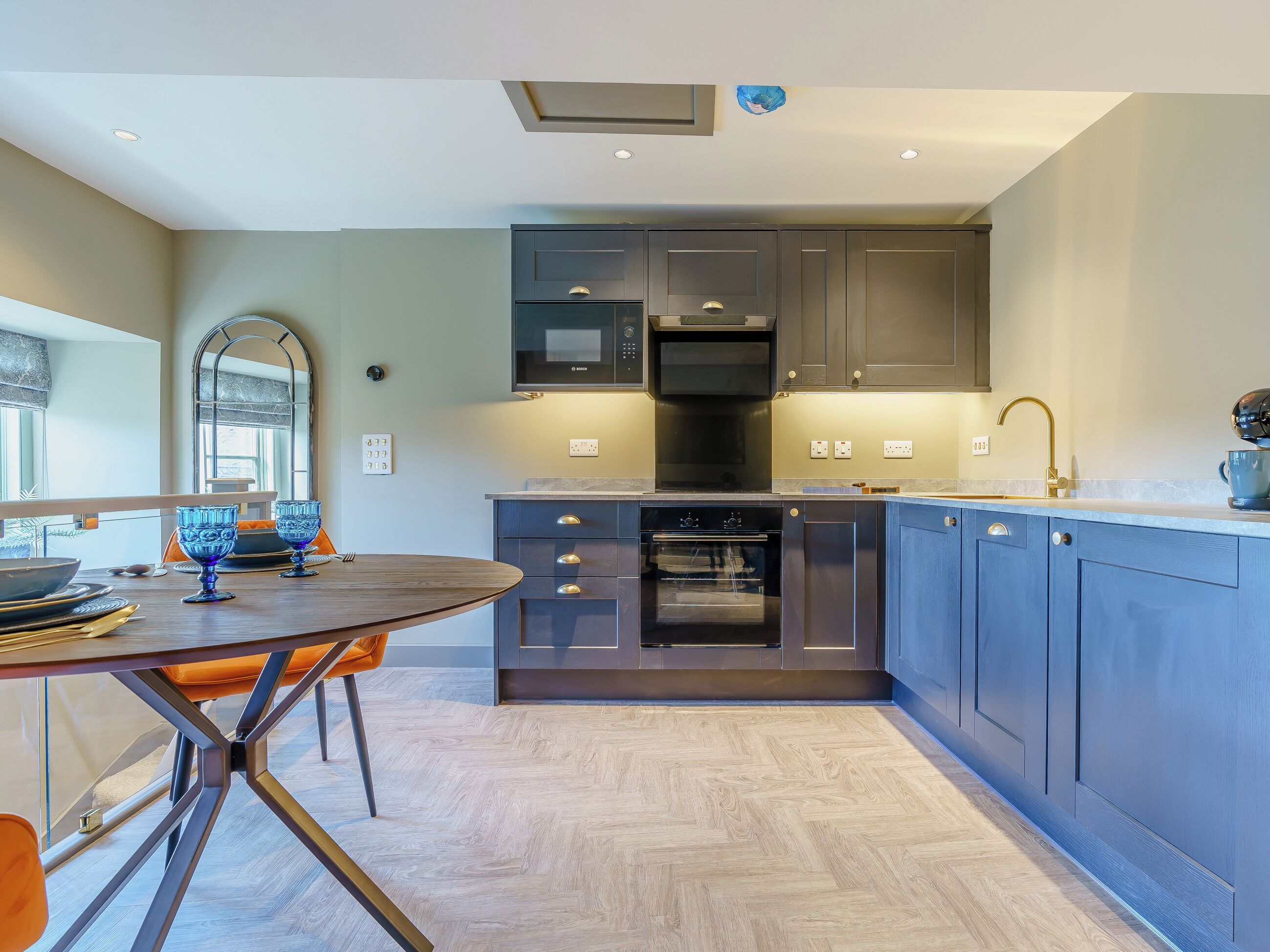 A modern kitchen with dark cabinetry, a built-in oven, and a dining area with a round table and orange chairs.