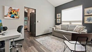 Uma sala de estar moderna com um sofá cinza, uma mesa de centro e uma escrivaninha com um computador. Há quadros nas paredes e uma janela com persianas.