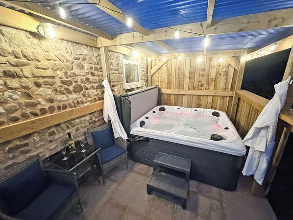 A stone-walled room with a wooden ceiling, a hot tub, a small table with bottles, and a bench.