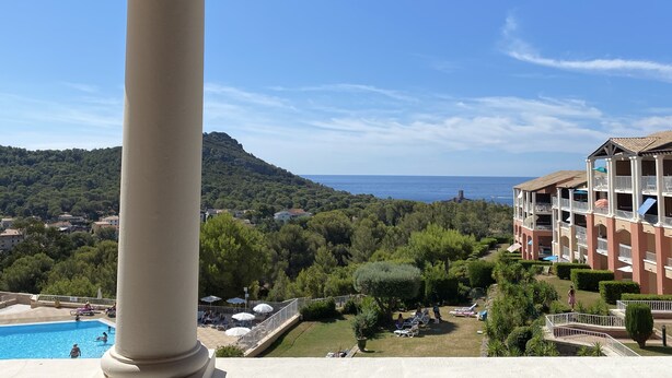 A view of a pool area with lounge chairs and umbrellas, a large white column, and a building with balconies overlooking a mountainous landscape. - Ideally located 4-person apartment with exceptional view (Saint-Raphaël)