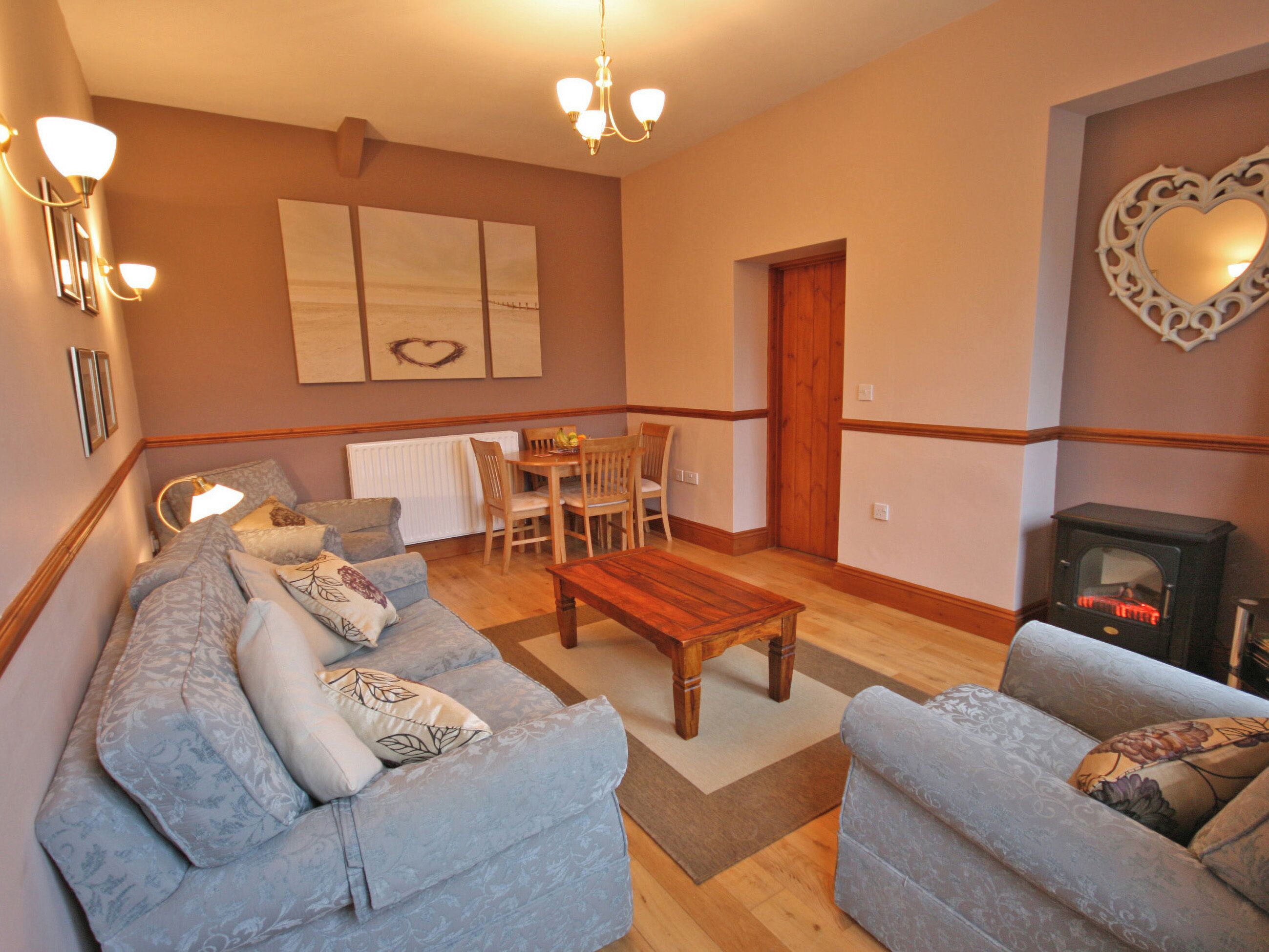 A living room with a sofa, armchair, wooden coffee table, and a fireplace. There is a dining area with a table and chairs, and artwork on the wall.
