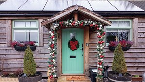 Una cabaña de madera con puerta verde, decorada con adornos navideños y un adorno de corona. Hay dos plantas en macetas a cada lado de la puerta.