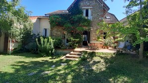 Una casa de piedra con tejado de tejas, un pequeño jardín con una mesa y sillas de madera, y un cielo azul despejado.