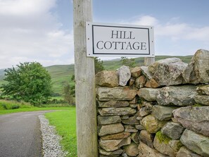 Un panneau indiquant l’emplacement de « Hill Cottage », un mur de pierres et une route de campagne.