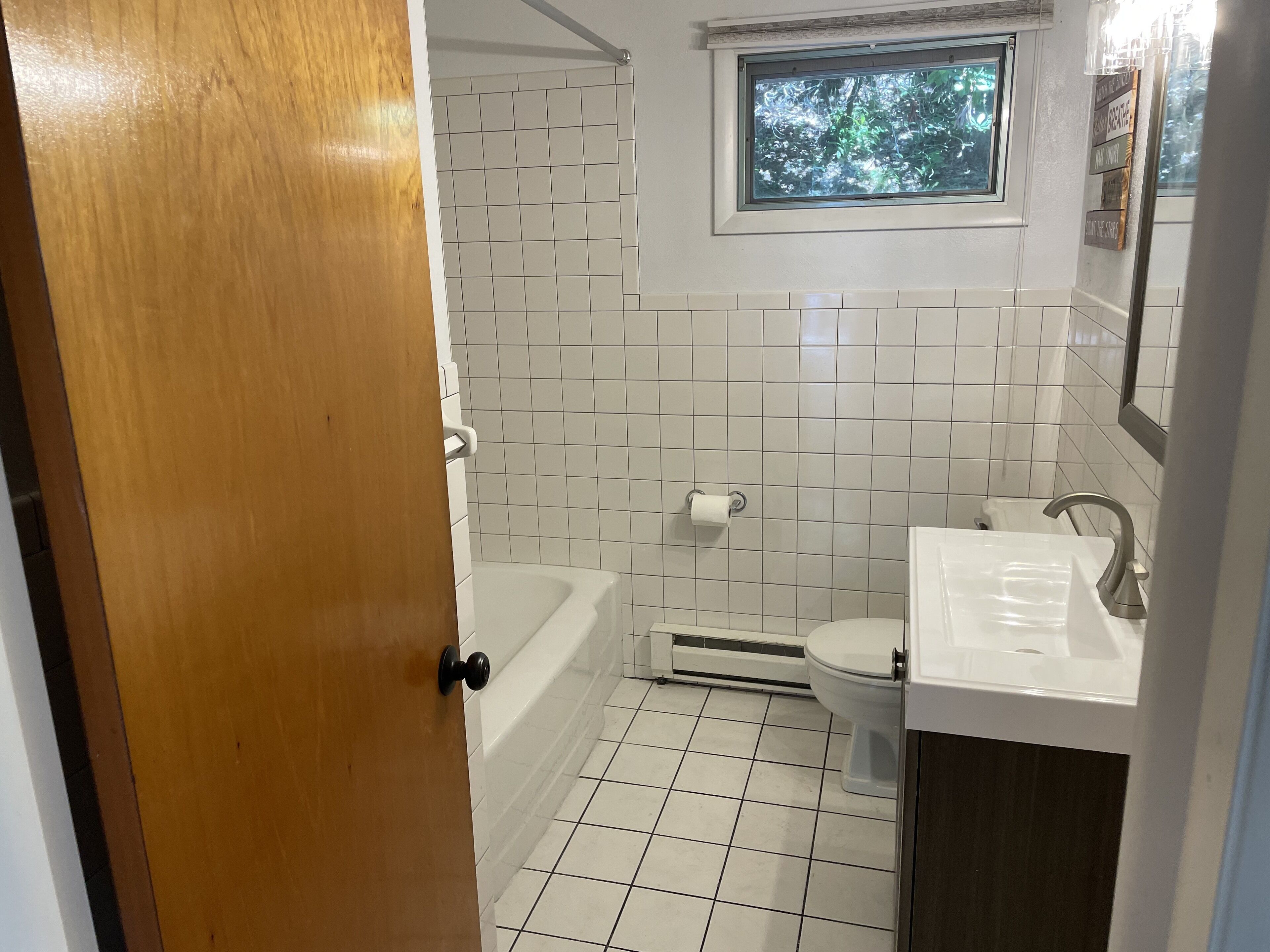 Un baño con bañera, inodoro y lavabo, con azulejos blancos y una ventana sobre el lavabo.