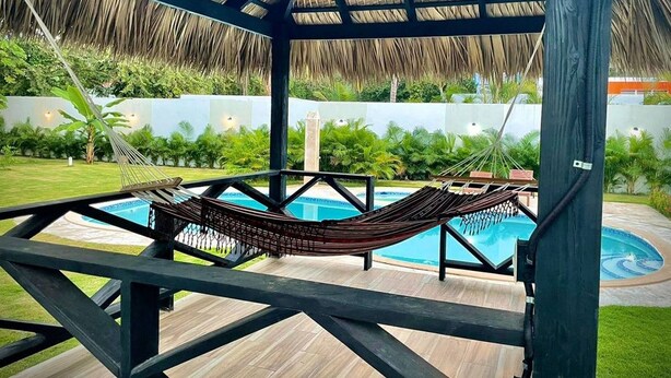 Un hamac sous une structure de toit en chaume, près d’une piscine avec une terrasse en bois et une végétation luxuriante en arrière-plan.