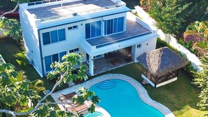 Une maison moderne à deux étages, dotée d’une piscine et d’une structure surmontée d’un toit de chaume.