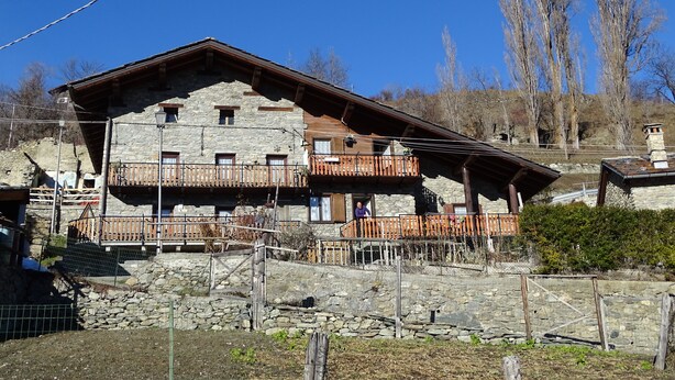 A stone building with wooden balconies and a sloped roof, surrounded by trees and a clear sky.