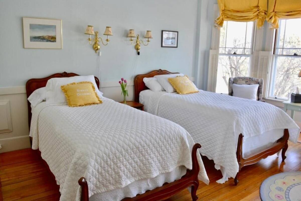 Dos camas de madera con colchas blancas y almohadas amarillas, un jarrón con flores y un cuadro en la pared.