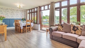 A dining area with a wooden table set for four, a sofa with patterned cushions, and large windows with curtains. - 3 bedroom accommodation in Beccles (Beccles)