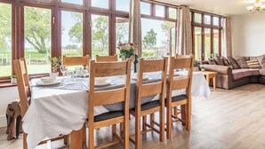 A dining area with wooden chairs and a table set for four, large windows with curtains, a chandelier, and a sofa in the background. - 3 bedroom accommodation in Beccles (Beccles)
