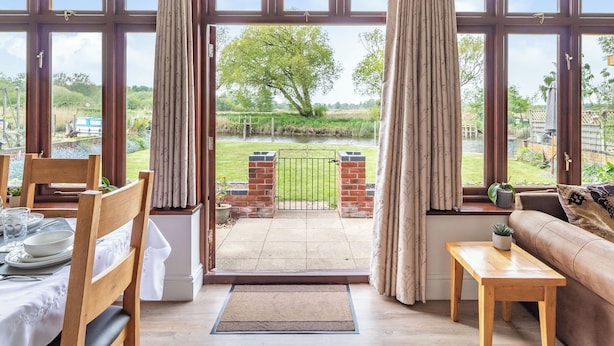 A conservatory with a view of a rural landscape, a dining area with a table set for two, and a sofa. - 3 bedroom accommodation in Beccles (Beccles)
