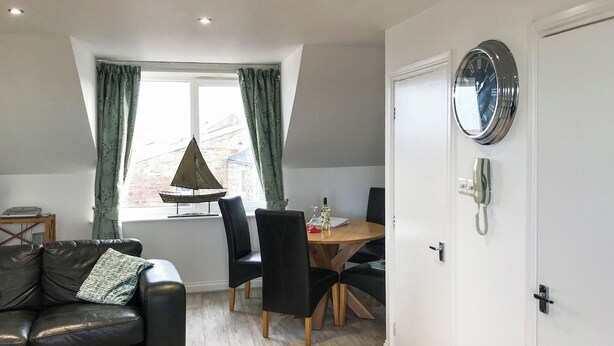 A modern living room with a black leather sofa, a dining table with chairs, a small sailboat decoration, and a wall-mounted telephone. - 2 bedroom accommodation in Seahouses (Seahouses)