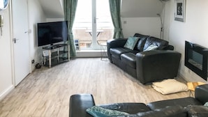 A living room with a black leather sofa, a television on a stand, and a balcony with curtains. - 2 bedroom accommodation in Seahouses (Seahouses)
