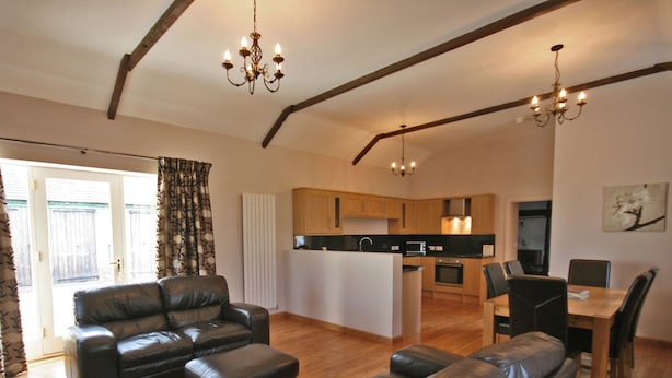 A modern living room with a leather sofa, a dining area with a wooden table and chairs, and a kitchen with wooden cabinets and a central island.