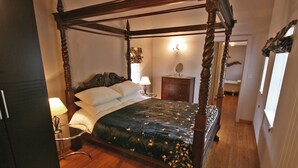 A four-poster bed with a dark floral bedspread, a bedside table with a lamp, and a mirror on the wall.
